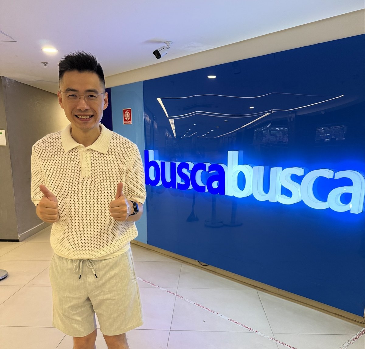 Alex Ye fazendo joinha com as duas mãos em frente à parede azul do Busca Busca